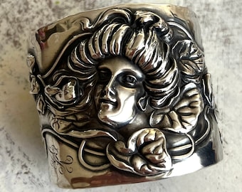 May include: A silver cuff bracelet with an ornate design featuring a woman's face surrounded by leaves and flowers.