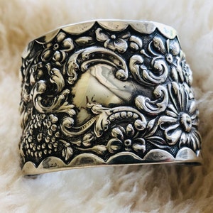 May include: A silver cuff bracelet featuring an elaborate floral design. The bracelet is wide and has a rounded shape. The design is raised and detailed, with flowers, leaves, and swirls.