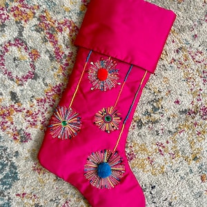 Puede incluir: Una media de Navidad rosa con un diseño acolchado y tres adornos bordados con cuentas y pompones de colores.