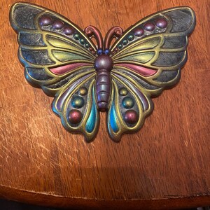Puede incluir: Un adorno de pared con forma de mariposa con detalles iridiscentes en dorado, azul, verde y morado. La mariposa tiene una superficie texturizada y está hecha de metal.