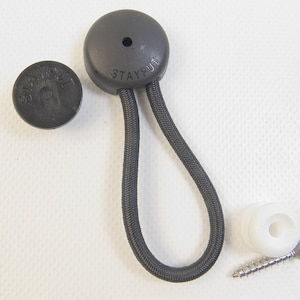May include: A black Stayput fastener featuring a circular plastic cap with a central hole and an attached elastic cord loop. The set includes an additional black circular cap, a white plastic mounting base, and a single metal screw for installation.