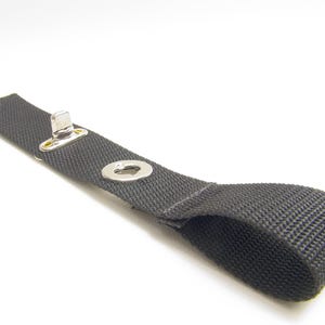 May include: A black strap with a metal loop and a metal button. The strap is made of a woven fabric.