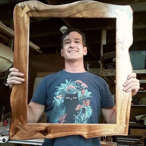 May include: A person holds a large, natural wood picture frame with a live edge. The frame is made of dark brown wood with a natural grain pattern. The person is wearing a blue t-shirt with a tropical print.