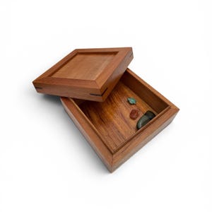 May include: A square wooden box with a removable lid, featuring a warm brown wood grain finish and dark wood corner accents. Inside the open box, there are three small, smooth, polished stones in shades of green, red, and dark gray.