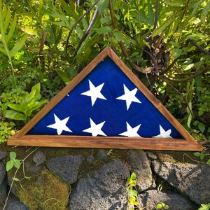 May include: A wooden flag display case with a folded American flag inside. The case is a triangular shape with a dark wood frame and a blue flag with white stars.