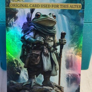 Pode incluir: Uma criatura verde semelhante a um sapo vestindo uma túnica marrom e carregando um cajado está em pé em uma rocha na frente de uma cachoeira. O cartão diz "ORIGINAL CARD USED FOR THIS ALTER" e "0/5".