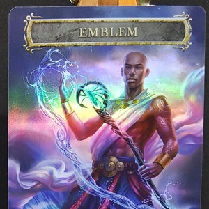 May include: A Magic: The Gathering emblem card with a blue, purple, and pink background. The card features a man with dark skin, holding a staff with a green orb at the top. The card text reads: "You may activate loyalty abilities of planeswalkers you control on any player's turn any time you could cast an instant."