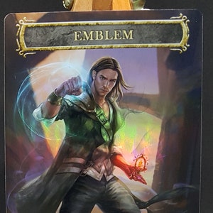 May include: A Magic: The Gathering card with a green, black, and red color scheme. The card features a man with long brown hair and a green jacket. The card text reads "EMBLEM" and "Whenever you cast a spell that targets one or more permanents, gain control of those permanents."