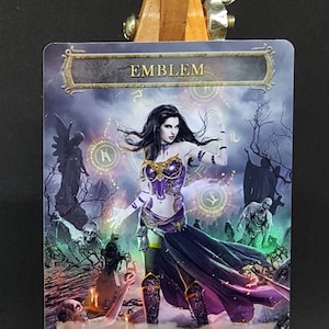 May include: A Magic: The Gathering card with a purple and gold emblem featuring a woman with long black hair and a purple and gold outfit. The text on the card reads "EMBLEM" and "Whenever a creature dies, return it to the battlefield under your control at the beginning of the next end step."