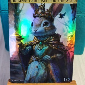 May include: A fantasy art card featuring a regal rabbit wearing a crown and ornate clothing. The card has a holographic effect and the text "ORIGINAL CARD USED FOR THIS ALTER" is visible. The card is propped up on a small wooden easel.