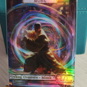 Może przedstawiać: Holograficzna karta do gry przedstawiająca postać mnicha z promienną aurą. Tekst na karcie brzmi "Monk" i "Token Creature - Monk 1/1 Prowess".