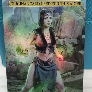 May include: A fantasy art card featuring a woman in warrior attire, holding a skull and a knife. The card has a gold banner that reads "ORIGINAL CARD USED FOR THIS ALTER". The background is a rocky landscape with green and yellow magical effects.
