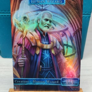 Könnte beinhalten: Eine Fantasy-Sammelkarte mit einem menschlichen Zauberer mit weißem Bart und einem Stab. Der Zauberer trägt eine lila und goldene Robe mit einem blauen Gürtel. Die Karte hat einen blauen Rand und den Text "HUMAN WIZARD" in weißen Buchstaben. Die Karte enthält auch den Text "Creature - Human Wizard" und "1/1" in schwarzen Buchstaben.