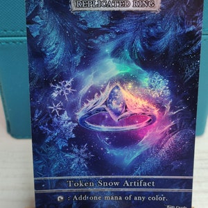 May include: A blue and purple fantasy-themed card with a silver border. The card features a ring with a large gemstone in the center. The card text reads "Replicated Ring" and "Token Snow Artifact". The card also has a mana symbol and the text "Add one mana of any color."