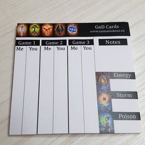 May include: A white card with a grid for tracking game scores. The card has a black header with the text "GnD Cards" and "www.customtokens.eu". The card is divided into three sections labelled "Game 1", "Game 2", and "Game 3". Each section has two columns labelled "Me" and "You". The card also has a section labelled "Notes" and a column on the right side with the text "Energy", "Storm", and "Poison".