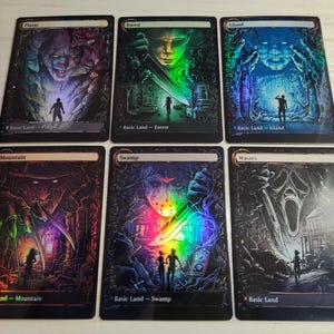 Op de afbeelding: Zes verzamelkaarten met horror-thema artwork. Elke kaart toont een andere scène met figuren en tekst. De kaarten zijn gelabeld met "Vlakte", "Bos", "Eiland", "Berg", "Moeras" en "Woestenij".