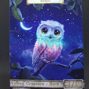 Könnte beinhalten: Eine weiße Eule mit schillernden Flügeln vor einem blauen Sternenhimmel. Die Eule sitzt auf einem Ast mit einem Halbmond im Hintergrund. Die Karte zeigt "Bird" und "Token Creature - Bird 1/1 Flying".