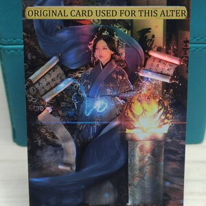 May include: A woman in a blue and black robe with a crown on her head stands in front of a glowing orb. She is surrounded by scrolls and a fire. The text "ORIGINAL CARD USED FOR THIS ALTER" is at the top of the image.