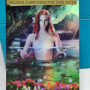 May include: A fantasy art card featuring a woman with red hair and a fishtail, holding a sword in a lake with lily pads. The card has a holographic finish and the text "ORIGINAL CARD USED FOR THIS ALTER" and "Art by Mary Neim".