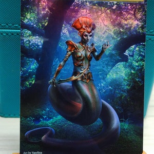 Könnte beinhalten: Eine veränderte Magic: The Gathering Karte mit einer Frau mit roten Haaren und einem Schlangen Schwanz. Die Karte ist vor dem Hintergrund eines Waldes mit einem blauen und violetten Schimmer platziert. Der Text "ORIGINAL CARD USED FOR THIS ALTER" ist in Gold oben auf der Karte gedruckt. Der Text "Art by Vasylina" ist in Weiß unten auf der Karte gedruckt.