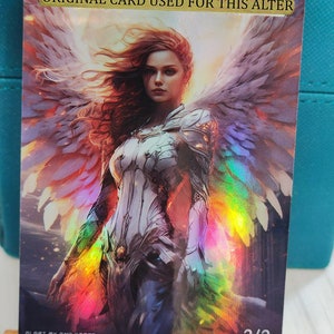 Pode incluir: Um cartão de arte de fantasia com uma mulher de cabelo vermelho longo e asas brancas. As asas têm um brilho iridescente arco-íris. O cartão tem o texto "ORIGINAL CARD USED FOR THIS ALTER" na parte superior e "2/2" na parte inferior.