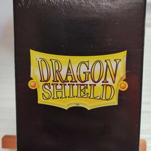 Peut inclure: Une bo&icirc;te noire avec un logo Dragon Shield dor&eacute; et le texte "100 Standard Size Sleeves".