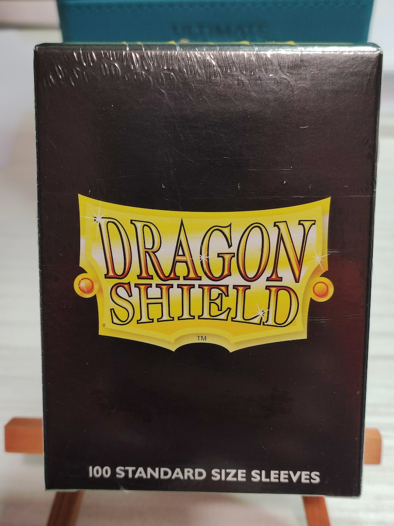Peut inclure: Une bo&icirc;te noire avec un logo Dragon Shield dor&eacute; et le texte "100 Standard Size Sleeves".