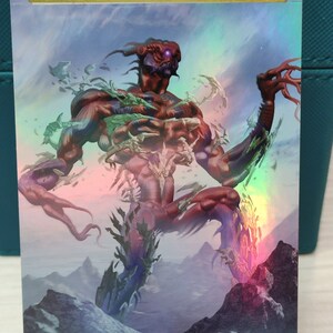 May include: A fantasy art card featuring a large, red, humanoid creature with a single, glowing purple eye. The creature is made up of many smaller, fragmented bodies, and is standing on a rocky mountain landscape. The card has a gold border with the text "ORIGINAL CARD USED FOR THIS ALTER" at the top.