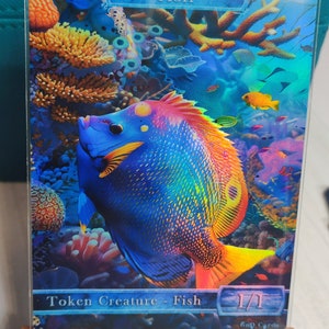 May include: A vibrant card featuring a colorful fish swimming in a coral reef. The fish has blue scales, yellow fins, and orange and red accents. The card reads "FISH" at the top and "Token Creature - Fish" at the bottom, with a 1/1 power/toughness.