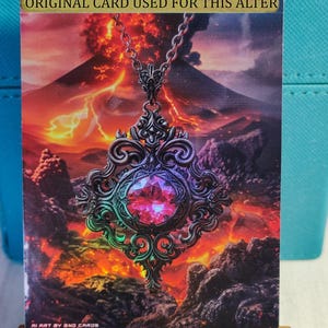 May include: A silver pendant with a large, faceted, pink gemstone hangs from a silver chain. The pendant is ornate and features intricate details. The background is a fiery orange and red landscape with a volcano erupting in the distance. The text "ORIGINAL CARD USED FOR THIS ALTER" is printed in gold at the top of the image. The text "AI ART BY GNO CARDS" is printed in white at the bottom of the image.