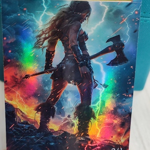 May include: A fantasy art card featuring a woman with long blonde hair holding a large axe. She is standing in a fiery landscape with lightning bolts in the background. The card has a holographic finish and the text "ORIGINAL CARD USED FOR THIS ALTER" at the top. The bottom right corner of the card has the number "2/1" printed on it.