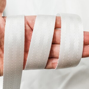 May include: White webbing strap with a textured surface. The strap is folded in half and held in a hand.
