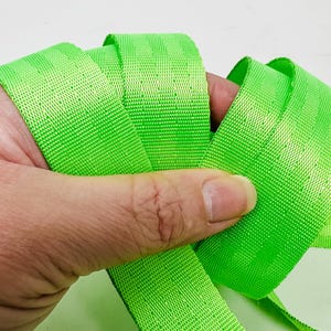May include: A bright green woven webbing strap, folded and held in a hand. The strap is made of a durable material and has a smooth, even texture.
