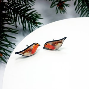May include: A pair of robin bird earrings. The earrings are made of resin and feature a realistic depiction of a robin with a red breast, brown back, and black eyes. The earrings are on a white background.
