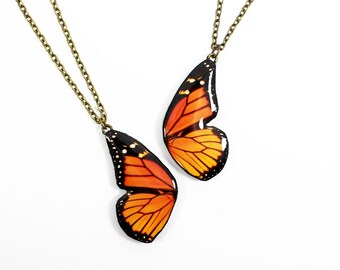 May include: Two necklaces featuring monarch butterfly wing pendants. Each pendant displays the iconic orange and black wing pattern with white spots, suspended from a thin, antique bronze-toned metal chain. The wings are shaped as individual halves.