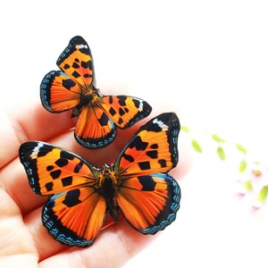 May include: Two orange and black butterflies with blue accents on a hand. The butterflies have a spotted pattern.
