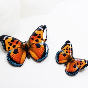 May include: Two orange and black butterfly brooches with blue accents. The larger brooch is on the left and the smaller brooch is on the right.