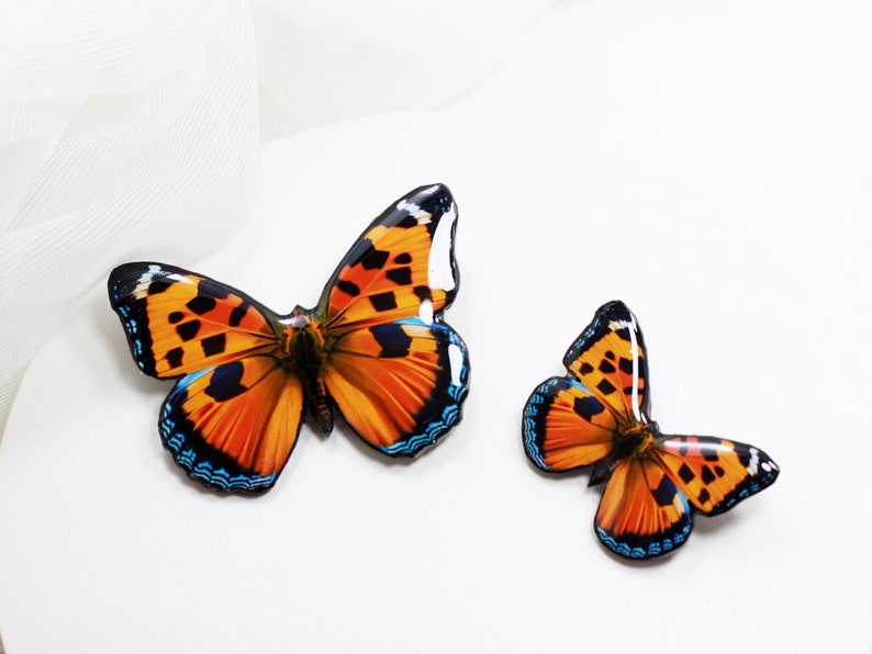 May include: Two orange and black butterfly brooches with blue accents. The larger brooch is on the left and the smaller brooch is on the right.