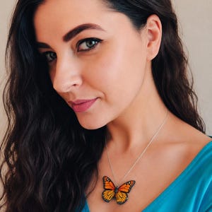 May include: A silver-toned necklace featuring a monarch butterfly pendant with vibrant orange, black, and white wing patterns. The butterfly is suspended from a delicate silver chain that rests against the collarbone, showcasing detailed wing markings.