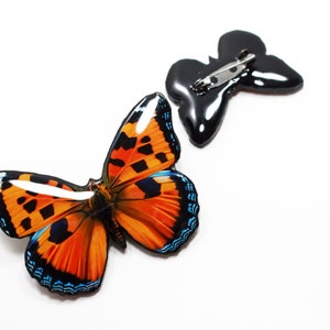 May include: A colorful butterfly brooch with orange, black, and blue wings. The brooch is attached to a black metal pin.