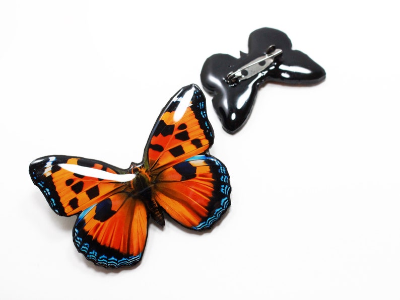 May include: A colorful butterfly brooch with orange, black, and blue wings. The brooch is attached to a black metal pin.