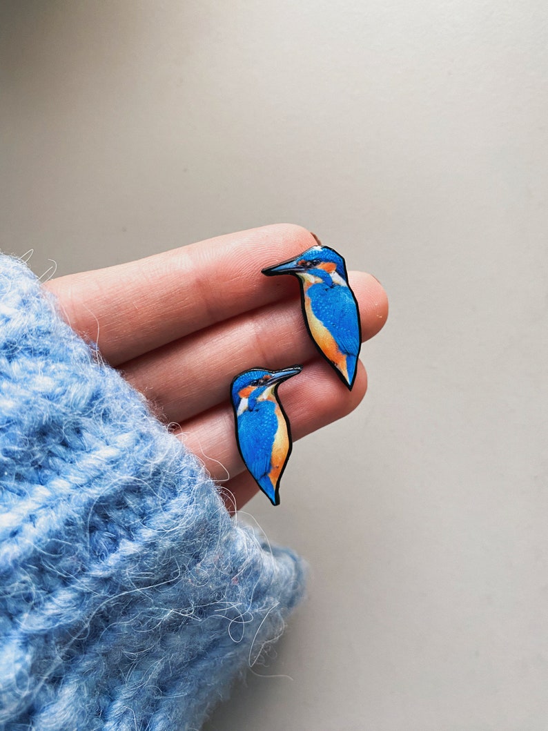 May include: A pair of blue and orange bird earrings. The birds are facing opposite directions and have black eyes and beaks.