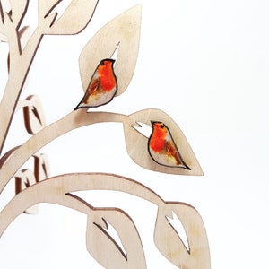 May include: A pair of robin bird earrings on a wooden tree display. The earrings are made of wood and have a realistic painted design.