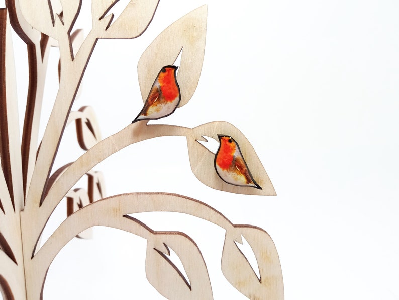 May include: A pair of robin bird earrings on a wooden tree display. The earrings are made of wood and have a realistic painted design.