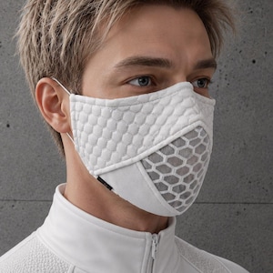 Op de afbeelding: Een wit gezichtsmasker met een gestructureerd zeshoekig patroon op het bovenste gedeelte en een rasterachtig ontwerp op het onderste gedeelte. Het masker wordt vastgezet met dunne oorlussen en heeft een klein zwart label op de zijnaad.