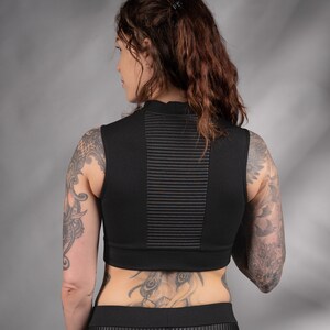 K&ouml;nnte beinhalten: Schwarzes &auml;rmelloses Crop-Top mit hohem Ausschnitt und gerippter Textur. Das Top wird mit einer schwarzen Hose getragen, die vorne eine lederartige Blende hat.