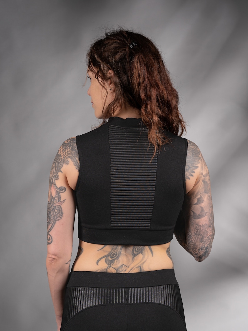 K&ouml;nnte beinhalten: Schwarzes &auml;rmelloses Crop-Top mit hohem Ausschnitt und gerippter Textur. Das Top wird mit einer schwarzen Hose getragen, die vorne eine lederartige Blende hat.