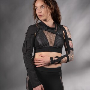 K&ouml;nnte beinhalten: Eine Frau tr&auml;gt ein schwarzes Mesh- und Leder-Crop-Top mit einem schwarzen Band &uuml;ber der Brust. Sie tr&auml;gt auch eine schwarze Hose mit Mesh-Eins&auml;tzen und schwarzen Armb&auml;ndern. Die Frau hat Tattoos auf ihrem Arm.