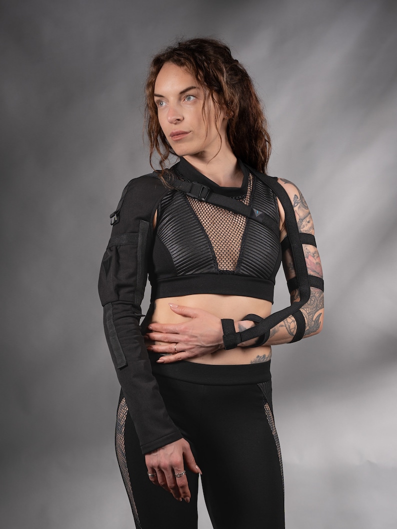 K&ouml;nnte beinhalten: Eine Frau tr&auml;gt ein schwarzes Mesh- und Leder-Crop-Top mit einem schwarzen Band &uuml;ber der Brust. Sie tr&auml;gt auch eine schwarze Hose mit Mesh-Eins&auml;tzen und schwarzen Armb&auml;ndern. Die Frau hat Tattoos auf ihrem Arm.