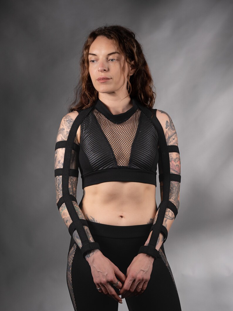 K&ouml;nnte beinhalten: Eine Frau tr&auml;gt ein schwarzes Crop-Top mit einem Mesh-Einsatz und schwarze Leggings. Sie hat Tattoos auf den Armen und tr&auml;gt schwarze Tr&auml;ger an den Armen.